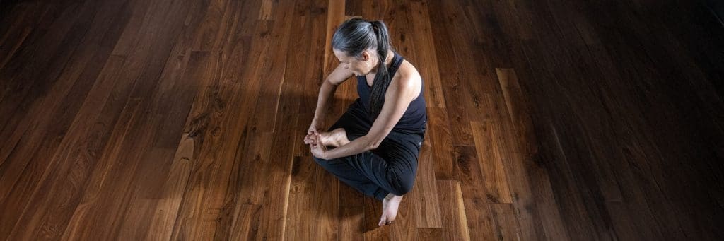 Alex Lambro in a seated Kaiut post, twisting at the waist and massaging her foot. She is seated on a wood floor with soft lighting